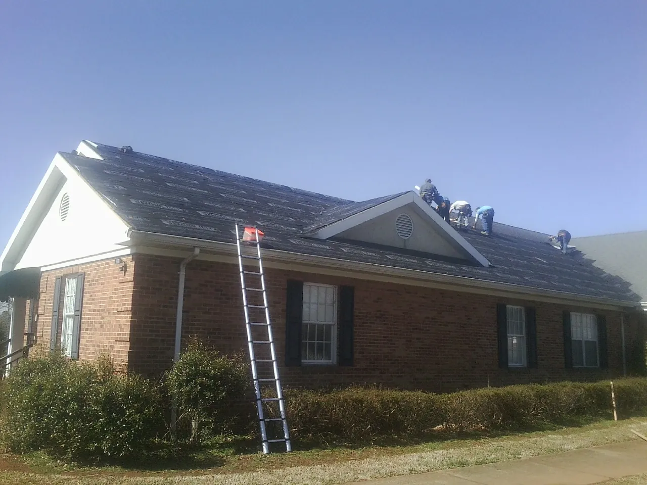 Crew tearing shingles off a brick ranch roof with a ladder set — Emergency Roof Repair work by our crew in Odenville