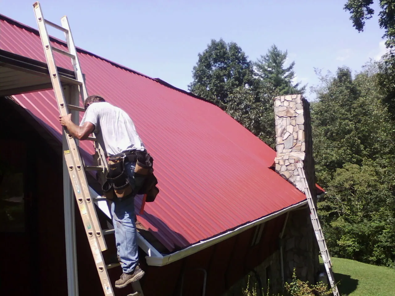 Roofer on a ladder beside a red metal roof and stone chimney — Soffit & Fascia Repair by Roofing Brothers in Odenville